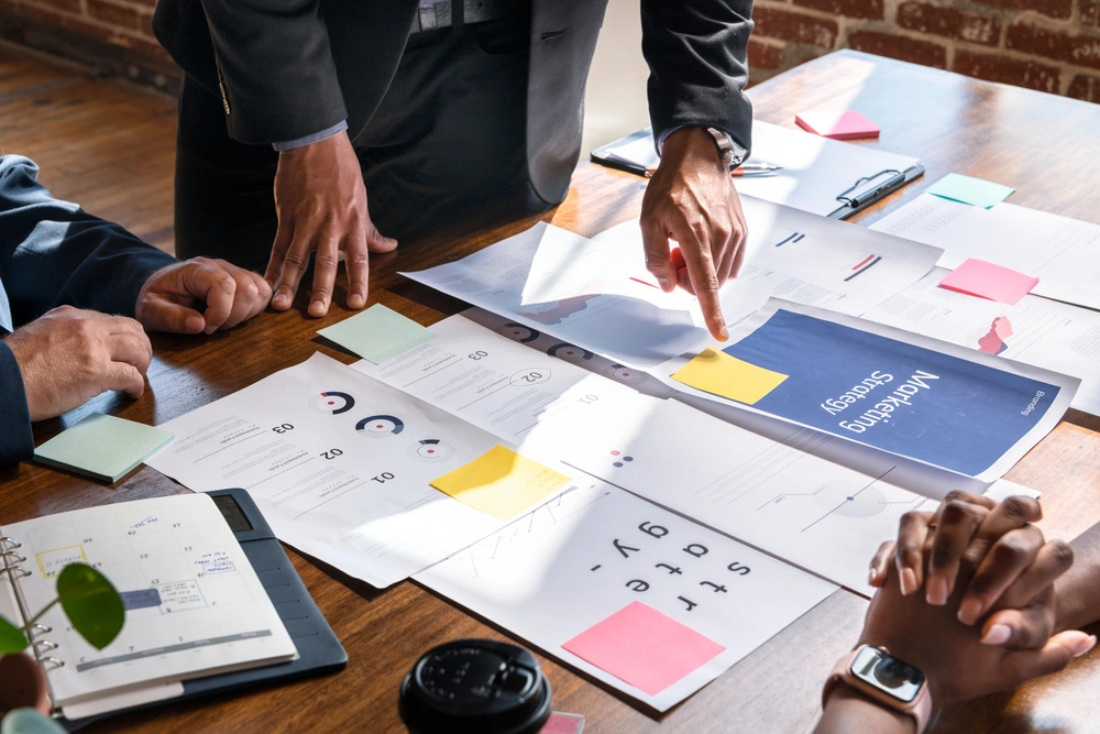 Business professionals reviewing printed marketing strategy documents on a wooden table, with charts, graphs, and sticky notes visible.