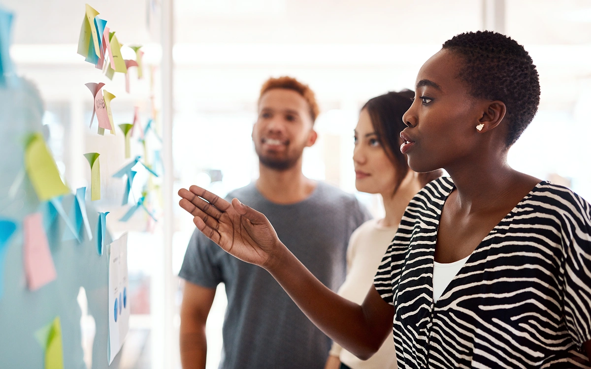 A diverse group of three colleagues collaborate in a modern office, standing at a whiteboard covered with colorful sticky notes. One person gestures toward the board while explaining an idea, as the others listen attentively.